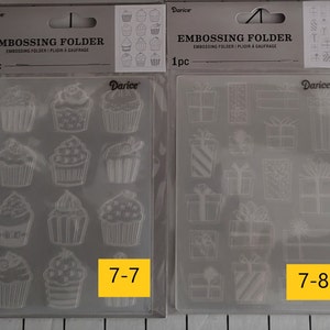 May include: Two clear plastic embossing folders for crafting. The folder on the left has a design of cupcakes. The folder on the right has a design of presents. Both folders have the number and text "7-7" and "7-8" printed on them.