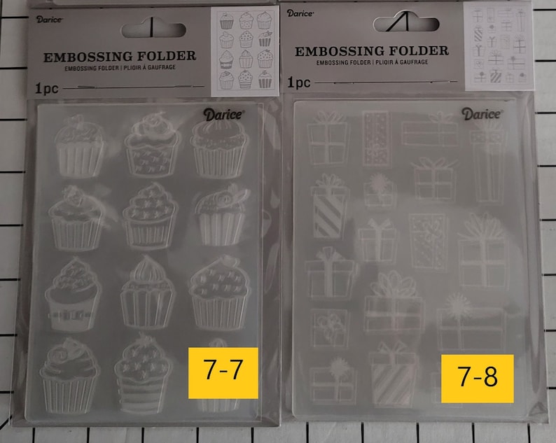 May include: Two clear plastic embossing folders for crafting. The folder on the left has a design of cupcakes. The folder on the right has a design of presents. Both folders have the number and text "7-7" and "7-8" printed on them.