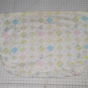 Puede incluir: Una sábana de cuna blanca ajustada con un patrón en amarillo pastel, azul y rosa. El patrón presenta arcoíris, estrellas y pequeños animales.