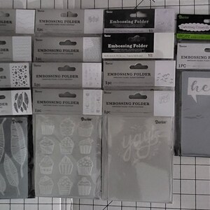 May include: A collection of 12 clear plastic embossing folders with various designs, including leaves, cupcakes, and the words "hello" and "you".