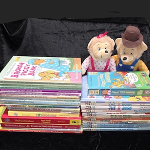 以下が含まれることがあります： 「Berenstain Bears」シリーズの子供向け書籍のコレクション。 「Broken Piggy Bank」や「Forget Their Manners」などのタイトルがあります。 2匹のぬいぐるみクマが本の後ろに配置されています。 本は黒い面に積み重ねられています。