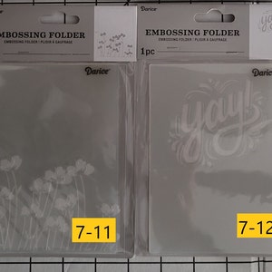 May include: Two clear plastic embossing folders for crafting. The folder on the left has a floral design and the text "7-11". The folder on the right has the word "yay!" in a stylized font and the text "7-12".