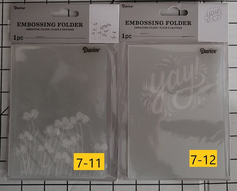 May include: Two clear plastic embossing folders for crafting. The folder on the left has a floral design and the text "7-11". The folder on the right has the word "yay!" in a stylized font and the text "7-12".