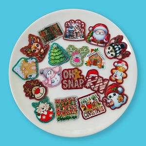 Pode incluir: Uma coleção de amuletos decorativos com tema natalino em um prato branco. Os amuletos apresentam vários designs, incluindo Papai Noel, renas, bonecos de gengibre e árvores de Natal. Os amuletos são coloridos com detalhes em glitter.
