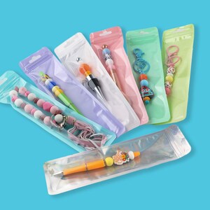 May include: A collection of colorful pens and keychains in clear plastic packaging. The pens feature decorative tops with beads and charms in various colors. The keychains have beaded accents and decorative elements. The background is a solid blue.