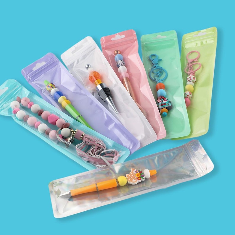May include: A collection of colorful pens and keychains in clear plastic packaging. The pens feature decorative tops with beads and charms in various colors. The keychains have beaded accents and decorative elements. The background is a solid blue.