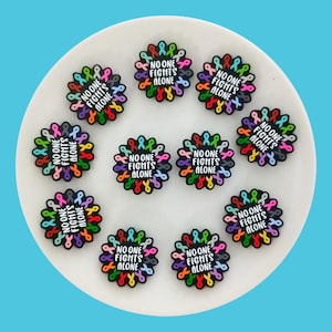 Op de afbeelding: Tien kleurrijke ronde pinnen met een cirkel van regenboog lintontwerpen en de tekst "No One Fights Alone" in wit.