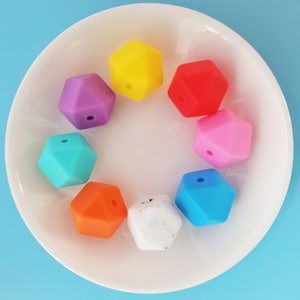 May include: Eight colorful silicone teething beads in various shapes, including a hexagon, a star, and a circle. The beads are arranged in a circle on a white plate.