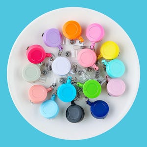 May include: Assortment of colorful retractable badge reels with a variety of colors including pink, blue, green, yellow, orange, and purple. The reels are on a white surface.