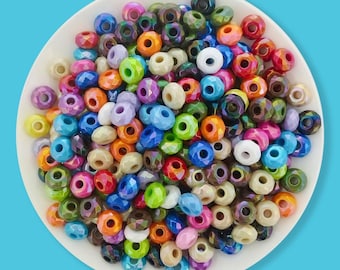 May include: A collection of colorful, faceted beads in various shades of blue, green, orange, pink, and white. The beads are round with a hole through the center, and are displayed on a white plate. The beads are likely used for jewelry making or crafts.