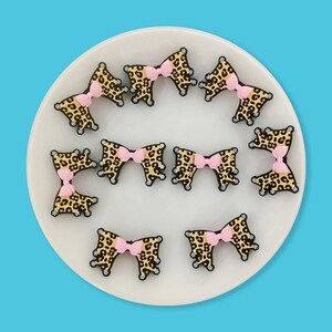 Op de afbeelding: Een verzameling strikvormige versieringen met luipaardprint en roze strikken, gerangschikt op een wit bord tegen een blauwe achtergrond. De strikken hebben een zwarte omtrek, voor knutselen of accessoires.