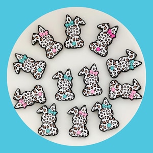 Op de afbeelding: Twaalf kleine, bruin-witte, luipaardprint konijnencharms met roze en blauwe strikjes. De charms zijn in een cirkel op een wit bord gerangschikt.
