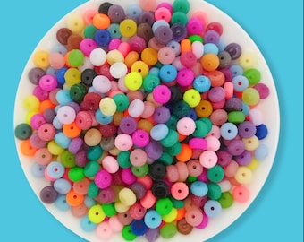May include: A white plate overflowing with a variety of colorful beads. The beads are in shades of pink, green, yellow, orange, blue, purple, and brown. Each bead has a hole through the center, suggesting they are for jewelry making or crafts.