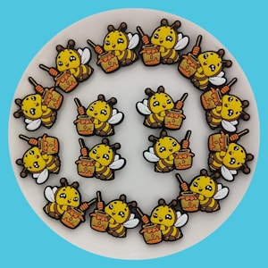 Puede incluir: Una colección de 15 abalorios de abeja amarillos y negros con tarros de miel. Cada abalorio de abeja tiene una linda cara de dibujos animados con ojos grandes y una sonrisa. Los abalorios están dispuestos en círculo sobre un plato blanco.