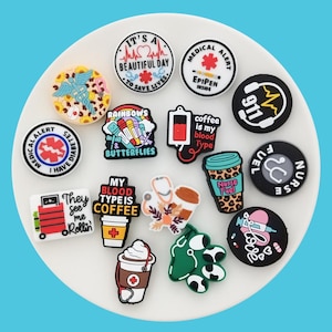Puede incluir: Una colección de 14 dijes de silicona redondos y cuadrados coloridos con varios diseños y textos, que incluyen "Es un hermoso día para salvar vidas", "El café es mi tipo de sangre", "Combustible de enfermería", "Mi tipo de sangre es café", "Me ven rodar", "Arcoíris y mariposas", "Alerta médica", "EpiPen dentro", "911", y "Tengo diabetes".