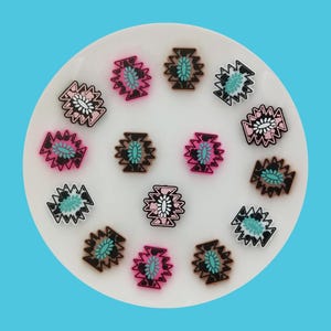 May include: A collection of decorative resin embellishments with a Southwestern design. The embellishments feature a geometric pattern in shades of pink, black, white, and turquoise. They are arranged on a white plate against a bright blue background.