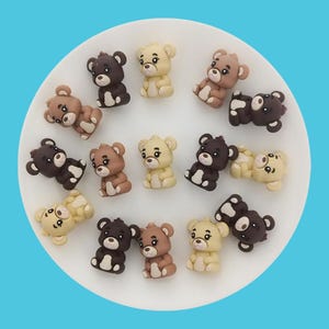 Puede incluir: Surtido de 15 pequeñas figuritas de ositos de peluche de color marrón, beige y crema. Los osos están todos sentados con las patas delante de ellos. Están dispuestos en círculo sobre un plato blanco.