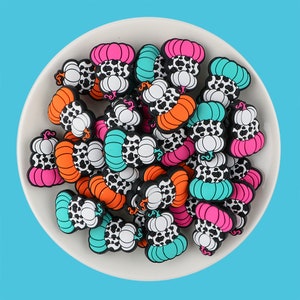 以下が含まれることがあります： 牛柄のデザインが施されたカラフルなカボチャ型のチャームのコレクション。チャームは柔らかく柔軟な素材で作られており、クラフトに秋の楽しさを加えるのに最適です。