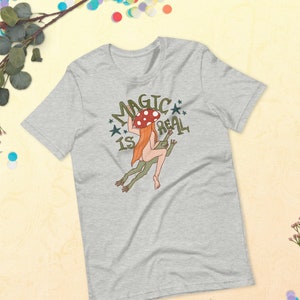 May include: A heather grey t-shirt with a graphic of a woman with red hair riding a frog. The woman is wearing a mushroom on her head. The text "Magic is Real" is written around the graphic.