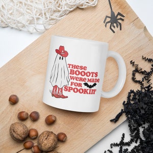 May include: White ceramic coffee mug with a ghost wearing red cowboy boots and a red cowboy hat. The text on the mug reads "These Boots Were Made For Spookin'" with a black bat graphic.