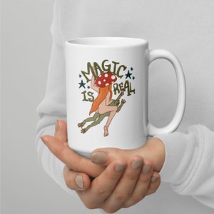 May include: White ceramic mug featuring a graphic of a woman with a mushroom on her head riding a frog. The text "Magic is Real" is printed on the mug.