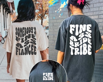 Op de afbeelding: Twee oversized T-shirts, één crème en één houtskool, met tekstafbeeldingen. Het crèmekleurige shirt zegt "Somebody's Fine Ass Fiancee" in zwart. Het houtskoolkleurige shirt zegt "Fine Ass Bride Tribe" in wit. Een kleinere afbeelding toont het houtskoolkleurige shirt met de tekst "Fine Ass Bride".