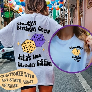 May include: Two white t-shirts with a graphic design. The first shirt has a purple and yellow dice design with the text "Sin-City Birthday Crew Julia's 30th Birthday Edition". The second shirt has a yellow smiley face with the text "Sin-City Birthday Crew".