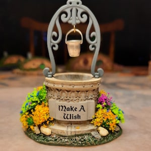 May include: A miniature stone-colored wishing well featuring a gray metal arch and a hanging bucket. The base is decorated with colorful orange, yellow, purple, and green floral accents. A plaque on the front displays the text Make A Wish.