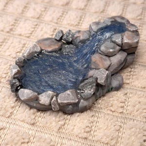 Pode incluir: Uma escultura decorativa de lago em miniatura feita de pedras cinzentas dispostas numa forma oval, com uma superfície de resina azul brilhante que imita a água a fluir para uma pequena piscina, criando um efeito realista de água.