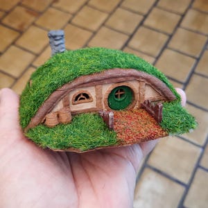 May include: A miniature hobbit house with a green grass roof, a round green door, and a small chimney. The house has a brown wooden frame and a stone path leading to the door. The model is held in a hand.