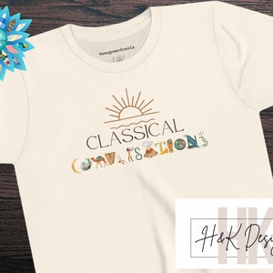 May include: A white t-shirt with a graphic design that says "Classical Conversations" with a sun and various icons. The text "Homegrown Grace Co." is printed on the top of the shirt. The bottom of the shirt has the text "H&K Designs" in a handwritten font.