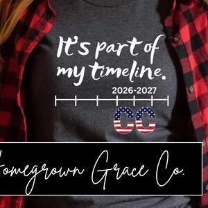 May include: Dark gray t-shirt with the text "It's part of my timeline" in white script. Below the text is the timeline graphic with the years 2026-2027. The letters "CC" are designed with the American flag. The shirt is worn with a red and black plaid shirt.