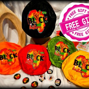 May include: Five fabric headbands in black, orange, red, green, and yellow. Each headband features a graphic of a raised fist and the words "I am Black History" with an outline of the continent of Africa. The headbands are on a white surface with several small round stickers with the words "Black History Month".