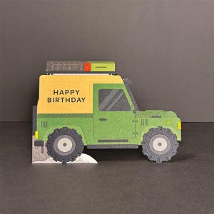 Könnte beinhalten: Grüne und gelbe Geburtstagskarte in Form eines Oldtimer-Geländewagens. Auf der Karte steht "HAPPY BIRTHDAY" auf der Seite. Das Fahrzeug hat große Reifen und einen Dachgepäckträger.