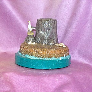 May include: A miniature scene of a tree stump with a bird perched on top, surrounded by seashells and sand, all set on a teal blue base.