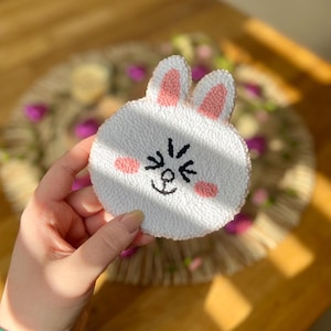 May include: A white plush bunny with pink ears and a black embroidered face. The bunny is smiling with closed eyes and has two pink cheeks.