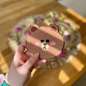 May include: A brown bear-shaped needle felted coaster with pink cheeks and a black nose.