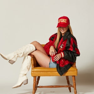 May include: A woman wearing a red and black jacket with the text "LFGKIC" on a red baseball cap, white cowboy boots, and denim shorts. She is sitting on a yellow chair and holding a red cup.