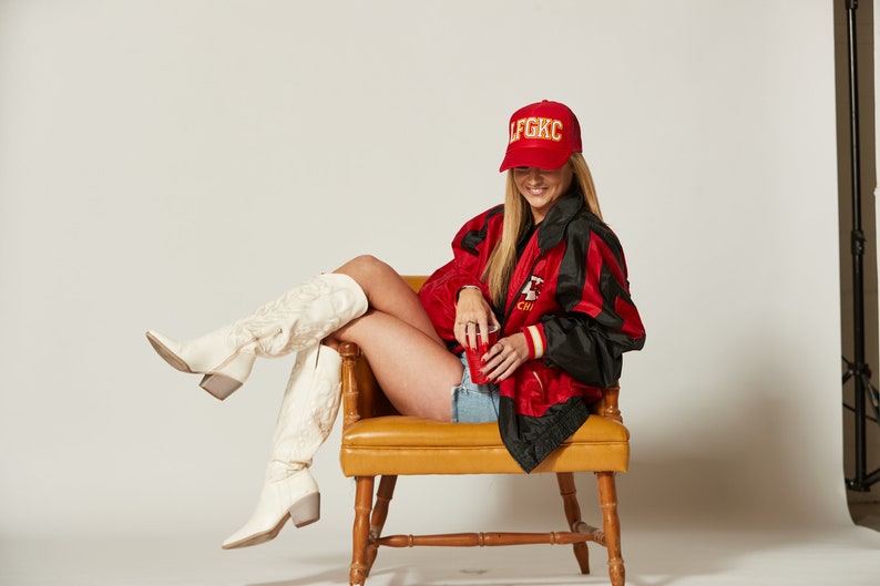 May include: A woman wearing a red and black jacket with the text "LFGKIC" on a red baseball cap, white cowboy boots, and denim shorts. She is sitting on a yellow chair and holding a red cup.