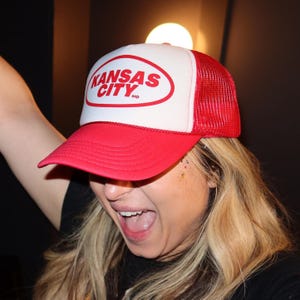 May include: A red and white trucker hat with a white oval patch that says "KANSAS CITY MO" in red letters.