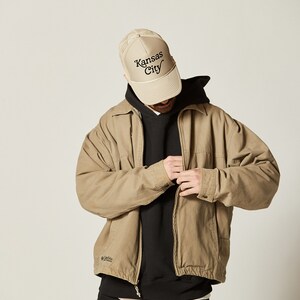 Pu&ograve; includere: Una giacca beige con chiusura a zip e un logo Columbia sul lato sinistro. La giacca &egrave; indossata sopra una felpa con cappuccio nera e jeans neri. La persona indossa un cappello da camionista in rete beige con il testo "Kansas City" ricamato in nero.