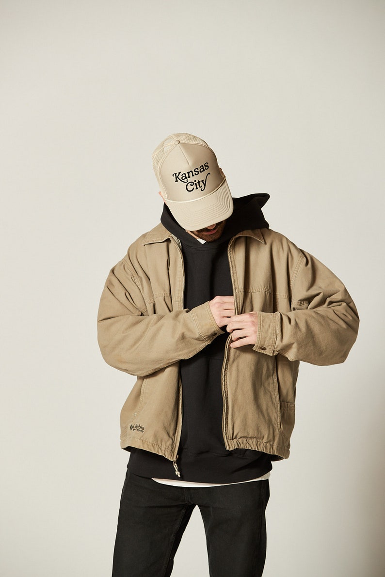 Pu&ograve; includere: Una giacca beige con chiusura a zip e un logo Columbia sul lato sinistro. La giacca &egrave; indossata sopra una felpa con cappuccio nera e jeans neri. La persona indossa un cappello da camionista in rete beige con il testo "Kansas City" ricamato in nero.