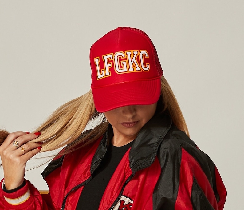 May include: A red trucker hat with a white "LFGK" logo in yellow outlined letters. The hat is worn by a person with long blonde hair and a red and black jacket.