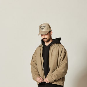 Pu&ograve; includere: Una persona che indossa una giacca beige con una felpa con cappuccio nera sotto. La persona indossa un cappello da baseball beige con il testo "Kansas City" ricamato sul davanti.