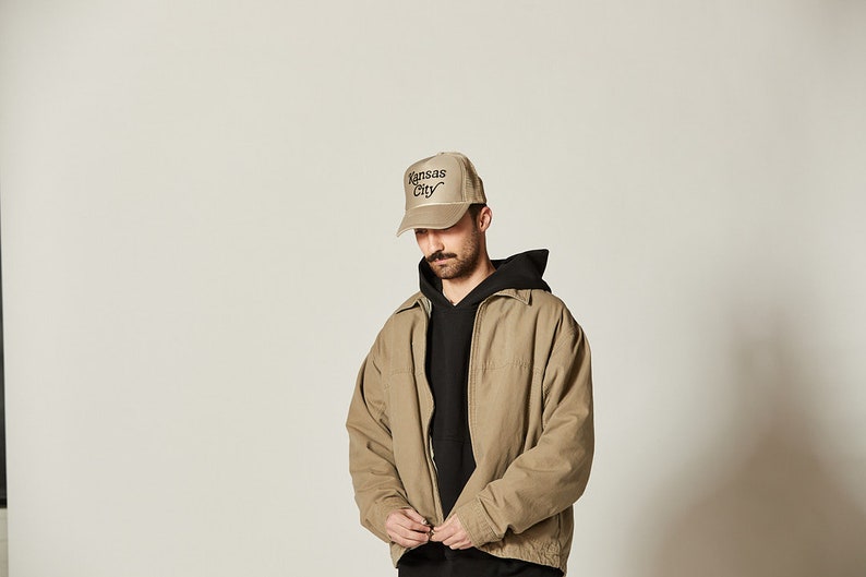Pu&ograve; includere: Una persona che indossa una giacca beige con una felpa con cappuccio nera sotto. La persona indossa un cappello da baseball beige con il testo "Kansas City" ricamato sul davanti.
