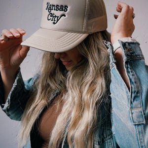 Pu&ograve; includere: Un cappello da camionista beige con un logo ricamato nero che dice "Kansas City".