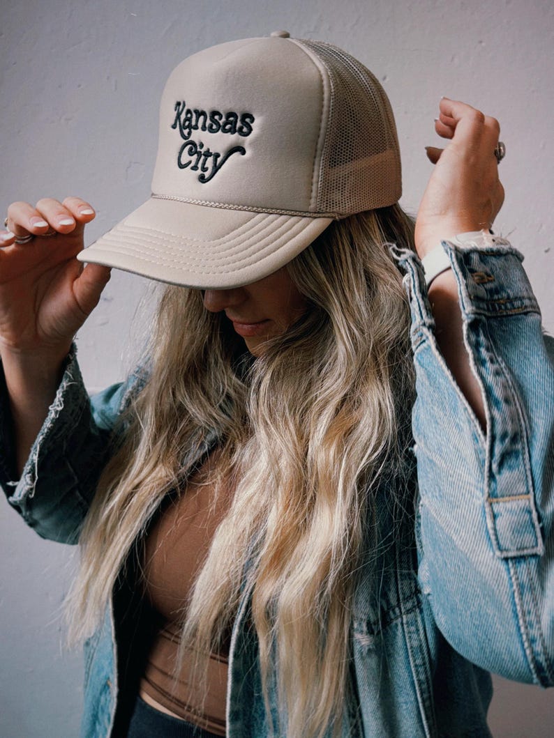 Pu&ograve; includere: Un cappello da camionista beige con un logo ricamato nero che dice "Kansas City".