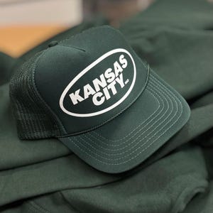 May include: A dark green trucker hat with a white oval patch that says "KANSAS CITY, MO".