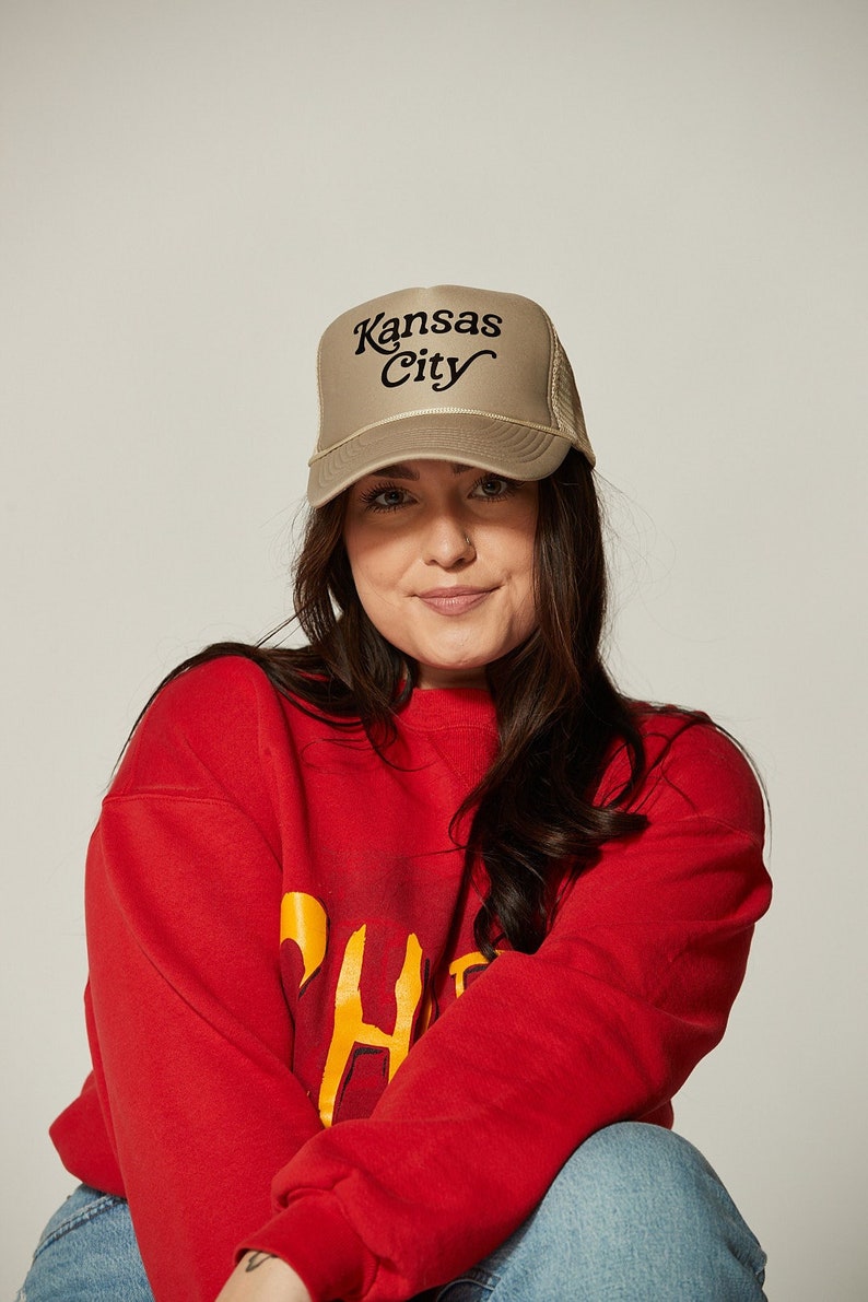 Pu&ograve; includere: Una donna indossa una felpa rossa con una grafica gialla e un cappello da camionista beige con il testo "Kansas City" in nero. Indossa jeans blu.