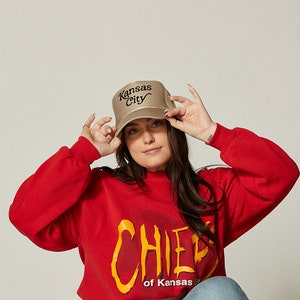 Pu&ograve; includere: Una donna indossa una felpa rossa con la scritta "CHIEF of Kansas" stampata e un cappello da camionista beige con "Kansas City" ricamato in nero.